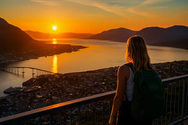 A blonde woman with a backpack stands on a viewing terrace above Tromso, looking at the city and the Tromsoysund Bridge in the golden light of the midnight sun; in the background, calm water, mountains, and the illuminated buildings of the city